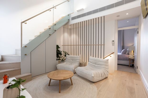 Bright contemporary living area with a light timber floor, sculptural cream lounge chairs, a round timber coffee table, and a pale textured rug. A glass-balustraded staircase with integrated under-stair storage rises beside a vertical timber slat feature wall, while a bedroom is visible through the open doorway. The space has high ceilings, recessed lighting, concealed air conditioning, and abundant natural light.