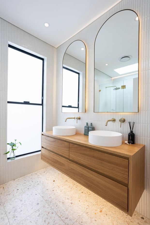 Bright contemporary bathroom featuring a wall-mounted double timber vanity with two white vessel basins, brushed brass wall-mounted taps, arched mirrors with dark trim, vertically ribbed light wall tiles, and a glass-enclosed shower visible in reflection. The floor is finished in pale terrazzo with multicoloured aggregate. A large frosted window and skylight provide abundant natural light.