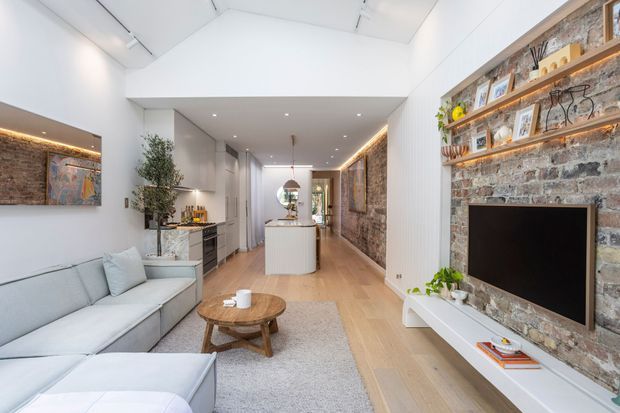 Bright open-plan contemporary living space with a white sectional sofa, timber coffee table, pale timber flooring and a large exposed-brick feature wall. Built-in timber shelving with integrated lighting surrounds a wall-mounted television, while a compact white kitchen and island extend down the room. The ceiling includes a high pitched section with skylights, creating an airy, elongated interior.