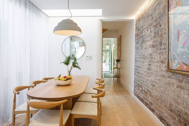 A bright dining area with a long rounded timber dining table and matching wooden armchairs, positioned beside full-height sheer curtains. A textured exposed brick feature wall runs along the right side with colourful framed artwork, while a sculptural pendant light hangs above the table. Timber flooring, a round wall mirror, indoor plant and a view through to the hallway create a warm, contemporary interior.