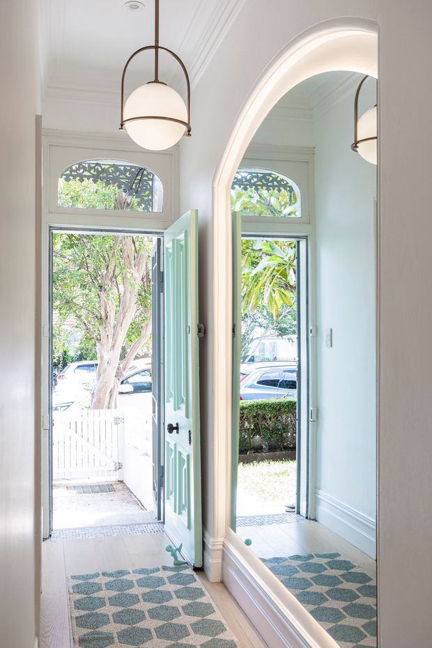 Bright, narrow residential entryway with a pale mint-green paneled front door, tall arched transom windows, ornate white mouldings, a large arched wall mirror, patterned blue-and-beige runner, and a suspended globe pendant light. The open doorway looks toward a small front path and garden, while the mirror reflects the entry and exterior greenery.