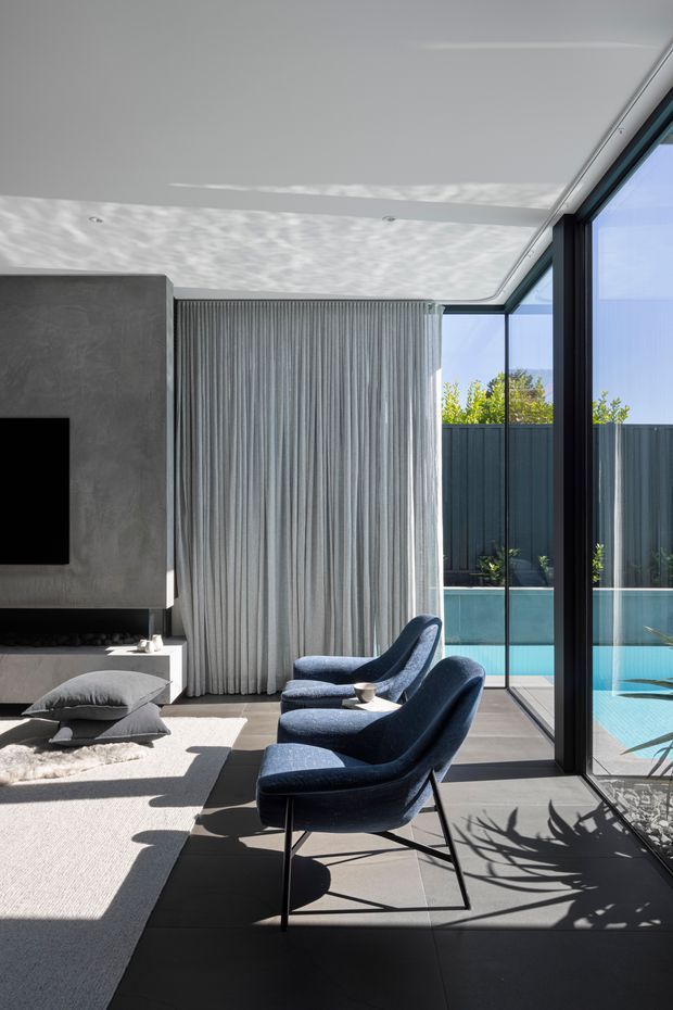 A refined contemporary living room with two deep blue upholstered lounge chairs facing a large wall-mounted television and dark concrete-look feature wall. Full-height glazing opens the room visually to a turquoise swimming pool, while sheer grey curtains, pale carpet, dark tiled flooring, and strong natural sunlight create a crisp indoor-outdoor composition.