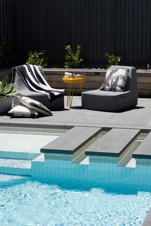 A contemporary poolside outdoor lounge with two dark upholstered armchairs, patterned cushions, a black-and-white throw, and a small yellow side table. The seating area sits beside a raised tiled swimming pool with narrow integrated stepping platforms. A dark vertically ribbed wall, timber planter bed, and green planting form the backdrop.