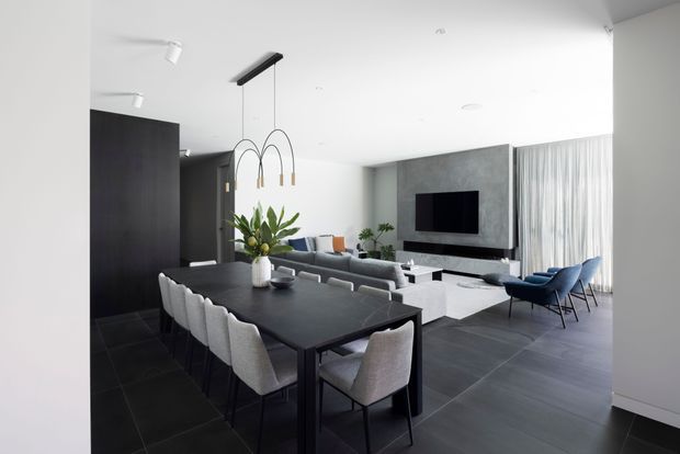 A refined open-plan living and dining area with a large black dining table and upholstered dining chairs in the foreground. The adjacent lounge includes a grey sofa, white coffee table, television mounted on a dark textured feature wall, and blue lounge chairs beside full-height sheer curtains. Dark tiled flooring, black joinery, minimalist lighting, and neutral furnishings create a contemporary, monochromatic interior.