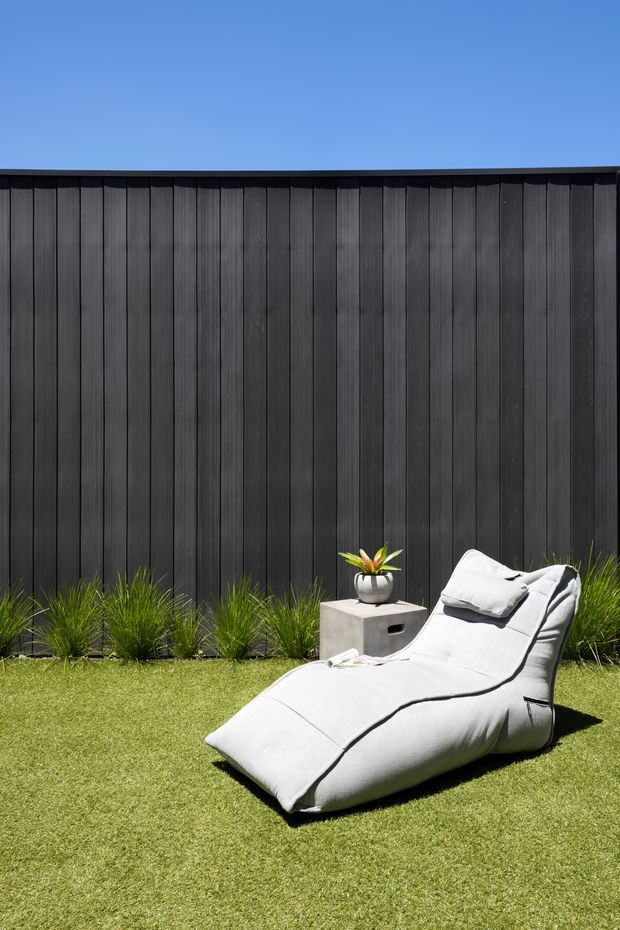 A minimalist outdoor relaxation area with a white cushioned chaise lounge on a neatly maintained lawn. Behind it is a tall dark vertical timber privacy fence, bordered by ornamental grasses. A small concrete side table with a potted plant sits beside the lounge. The scene is sunlit, contemporary, and carefully landscaped.