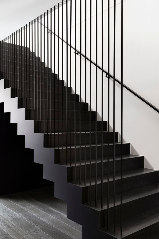 A minimalist interior stairway with a sculptural dark staircase, black vertical metal balusters, and a slim diagonal handrail against white walls. The stairs appear finished in dark timber or dark-toned material, contrasting with grey timber flooring and strong geometric shadows.