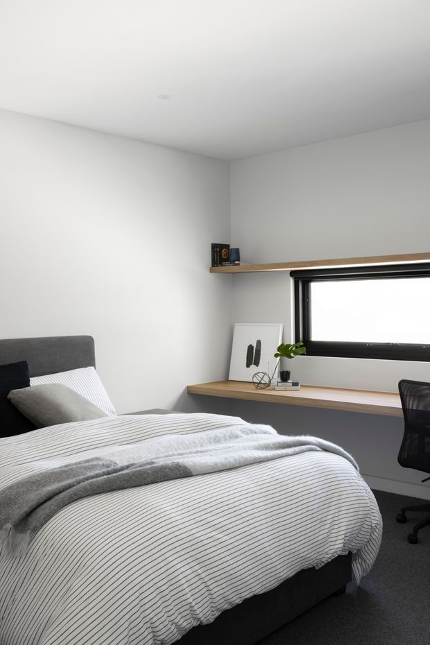 A compact, minimalist bedroom with a neatly made bed in white and fine grey stripes, a dark upholstered headboard, grey carpet, and white walls and ceiling. A black-framed horizontal window provides natural light, with a timber shelf and matching built-in desk beneath it. A simple office chair and sparse decorative objects complete the room.