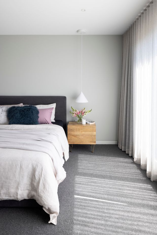 A bright, softly styled contemporary bedroom with a partially visible bed, dark upholstered headboard, layered white and pale pink bedding, and a deep blue accent cushion. A warm timber bedside chest with geometric drawer fronts sits beneath a simple white pendant light. Full-height sheer and grey curtains filter strong daylight onto the grey carpet, while the walls and ceiling are kept in restrained neutral tones.