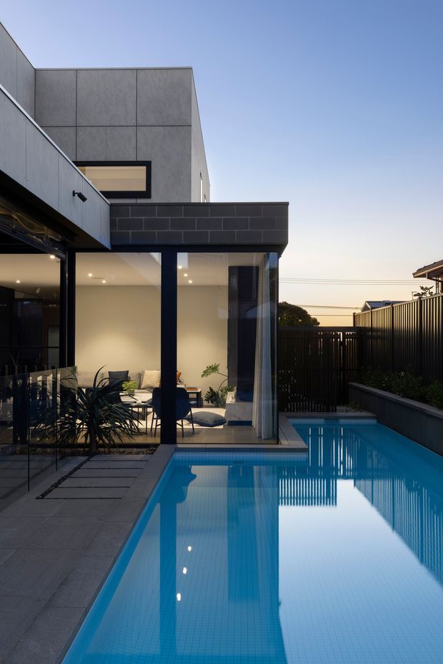 A dusk exterior view of a contemporary two-storey residence with crisp geometric forms, dark charcoal masonry, grey panel cladding, and extensive floor-to-ceiling glazing. The image looks across a narrow rectangular swimming pool toward a glazed living area, with a paved terrace, dark privacy fencing, and integrated outdoor planting. The pool and interior lights create strong reflections, while the clean lines and dark palette give the scene a refined modern character.