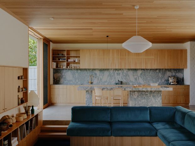 Open-plan contemporary living and kitchen space with extensive light timber cabinetry and a slatted timber ceiling. A deep blue sectional sofa occupies the foreground, facing a marble-look stone kitchen splashback and island with stools. Built-in shelving and joinery line the left wall, while glazed doors bring in natural light.
