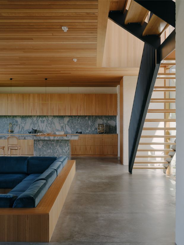 A contemporary open-plan interior combining a timber-lined kitchen and living area with a dramatic black steel stair. The kitchen features extensive warm timber cabinetry, a blue-grey veined stone splashback and island, and a polished concrete floor. A deep navy sectional sofa sits within a timber-framed built-in arrangement in the foreground. Open-riser timber stairs with black metal stringers lead upward beside a tall light-filled opening.