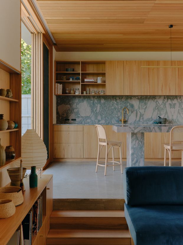 Warm, carefully detailed open-plan kitchen and living area with extensive light timber cabinetry and matching slatted ceiling. A dramatic blue-grey veined stone splashback and island surround the brass-toned kitchen tap. Open shelving displays ceramics and glassware, with two light woven bar stools at the island. Timber steps lead down toward a deep blue sofa in the foreground, while large glazing and vertical timber battens bring in leafy views and natural light.