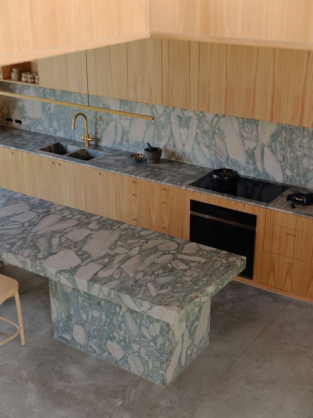 A contemporary kitchen with extensive green-and-white veined stone surfaces wrapping the island benchtop, waterfall sides, splashback, and rear benchtop. Light natural timber cabinetry runs along the walls, with integrated appliances, a black cooktop, undermount sink, and brass-toned gooseneck tap. The space has a polished yet natural material palette, concrete flooring, and a compact island with seating.