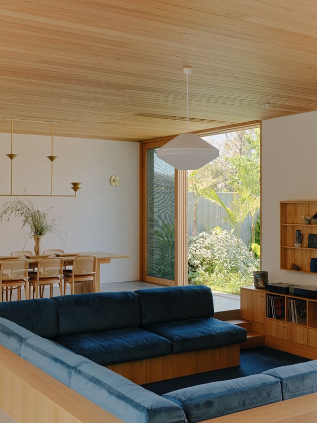 A warm, light-filled open-plan living and dining area with a deep navy-blue sectional sofa in the foreground, a timber-framed built-in entertainment unit on the right, and a timber dining table with woven-back chairs at the rear. The room features extensive pale timber lining across the ceiling, white textured walls, large glazed doors opening toward a leafy garden, sculptural pendant lighting, and mid-century-inspired built-in joinery. The composition emphasises the living room while showing the connected dining zone.