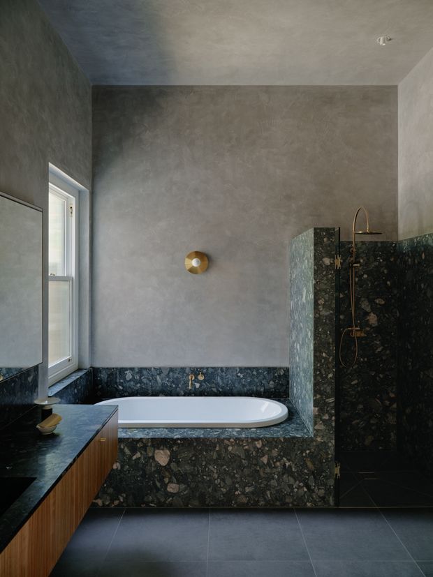 A dark, refined bathroom with a freestanding white bathtub set into a deep green-black stone surround, matching stone wall cladding around the shower and vanity. The room has smooth grey plaster walls and ceiling, a dark tiled floor, timber vanity cabinetry, a narrow window, and brushed brass shower and wall light fittings.