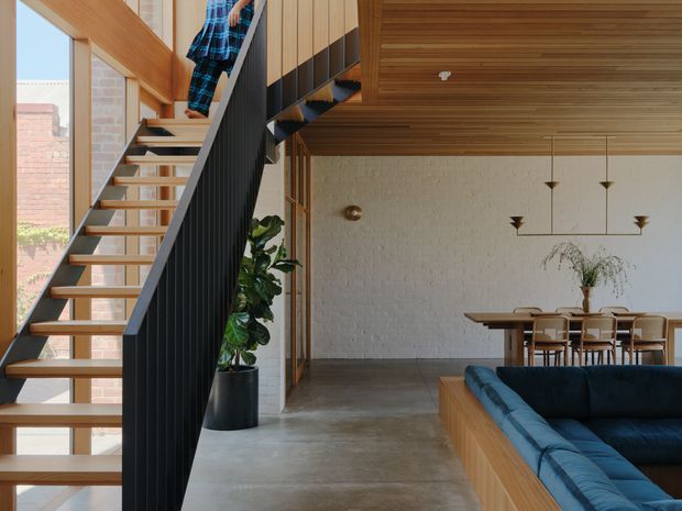 A bright contemporary open-plan interior viewed beside a sculptural staircase. The staircase has open timber treads, dark vertical balustrade screening and timber handrails, rising beside large windows. A dining area with a timber table and chairs sits against a light textured masonry wall, while a blue upholstered built-in or sectional sofa occupies the foreground. The room features extensive warm timber ceiling lining, exposed concrete flooring, minimalist pendant lighting and indoor planting.
