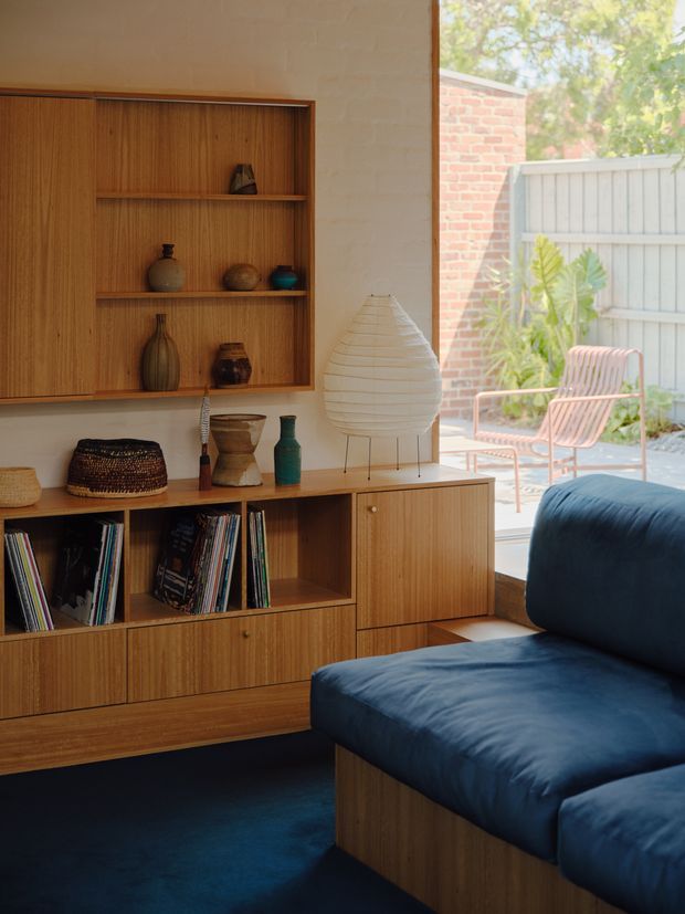 A warm, carefully styled living room featuring extensive built-in timber cabinetry with open shelving, drawers, and cupboards, displaying books and ceramics. A deep blue upholstered sectional sofa sits on a dark blue carpet beside a large sculptural paper floor lamp. Full-height glazing opens visually to a small paved courtyard with a timber slat chair, pale fence, and brick wall. The room has a strong mid-century-inspired aesthetic with natural timber, rounded forms, and warm daylight.