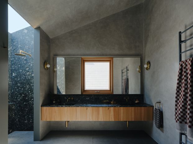A moody contemporary bathroom featuring a wide floating timber vanity with a dark stone benchtop, integrated twin basins, and a large wall mirror. Grey textured plaster walls and matching floor tiles create a monolithic feel. A separate shower area with dark natural stone wall cladding is visible at left, while heated towel rails and patterned towels occupy the right wall. A timber-framed window and brass wall sconces add warmth.