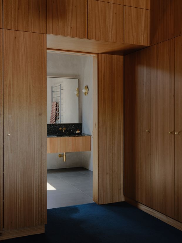 A refined walk-in wardrobe lined with full-height, warm timber veneer cabinetry and integrated flush doors. The wardrobe opens directly into a compact bathroom with a floating timber vanity, dark stone benchtop and splashback, wall mirror, brass tapware, and grey tiled floor. The foreground has a deep navy carpet, while the bathroom is softly lit with sculptural wall sconces.