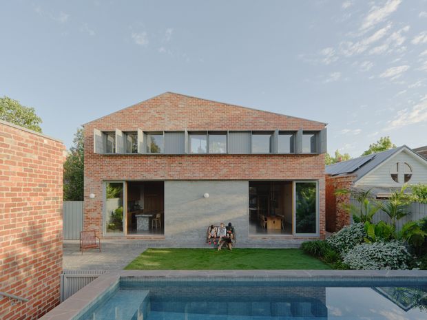 Rear exterior view of a contemporary two-storey brick residence enclosing a landscaped courtyard and swimming pool. The facade combines red brickwork, a broad grey concrete or masonry ground floor, extensive upper-level glazing, and a low pitched roof. A neighbouring gabled structure and garden planting are visible around the property.
