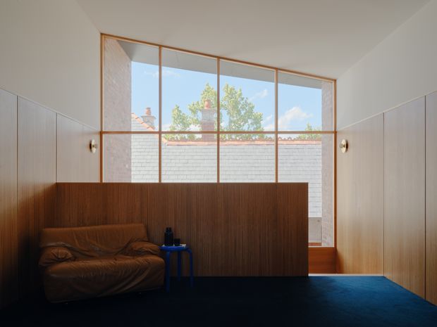 A compact, refined interior with a low brown leather sofa, dark blue carpet, and extensive pale timber wall panelling. A dark timber half-height partition separates the seating area from a large floor-to-ceiling window with warm timber framing, overlooking a brick boundary wall and greenery. The space has a restrained, gallery-like character with strong natural light and carefully composed geometric lines.