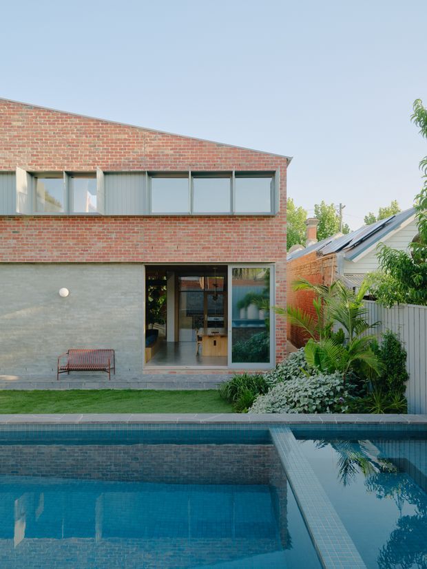 Exterior view of a contemporary two-storey brick house with a low-sloping roofline, broad horizontal glazing, and a textured grey ground-floor wall. The house opens onto a landscaped garden and swimming pool, with a smaller pitched-roof structure visible beside it.