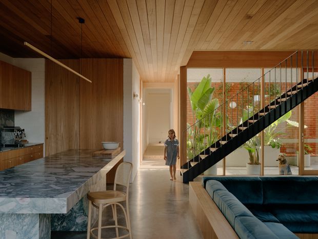 A warm, open-plan contemporary interior combining a kitchen, living area and stairway. The kitchen features timber cabinetry and a large polished stone island with stools, while a black metal open-riser staircase crosses in front of full-height glazing. Timber-lined ceilings, polished concrete flooring, brick walls and indoor planting create a refined, natural material palette.