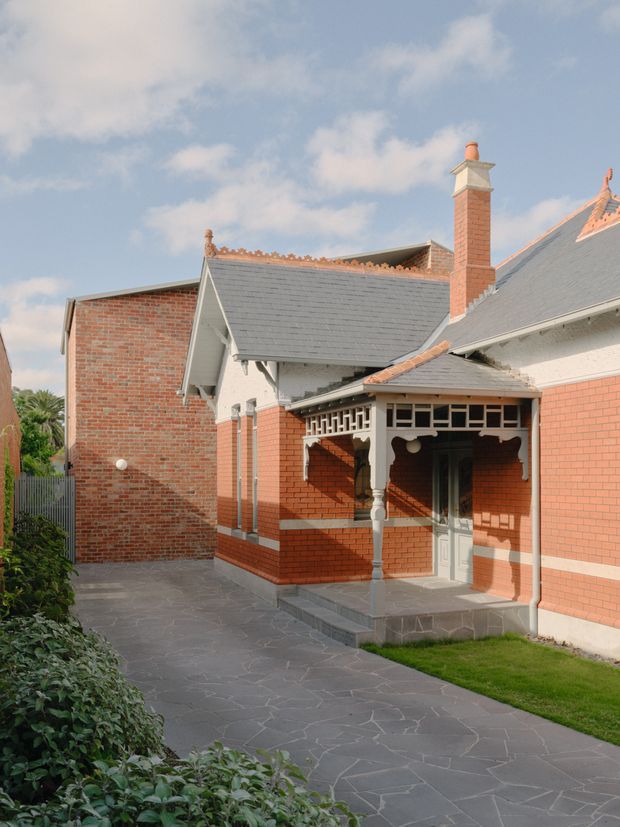 Exterior view of a heritage-style brick residence centered on a covered front entry porch. The building features red brick walls, grey slate-like pitched roofs, ornate timber fretwork and decorative gable detailing, a prominent brick chimney, and a paved path leading to the entrance. The courtyard-like approach includes low garden planting and lawn, under bright natural daylight.