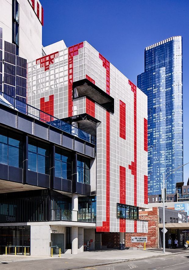 Street-level exterior view of a contemporary urban commercial and mixed-use building. The main facade features a white perforated or gridded envelope with bold red vertical and rectangular accents, projecting dark balconies or structural elements, and extensive dark glazing. A tall blue-glass high-rise and surrounding urban retail streetscape are visible in the background.