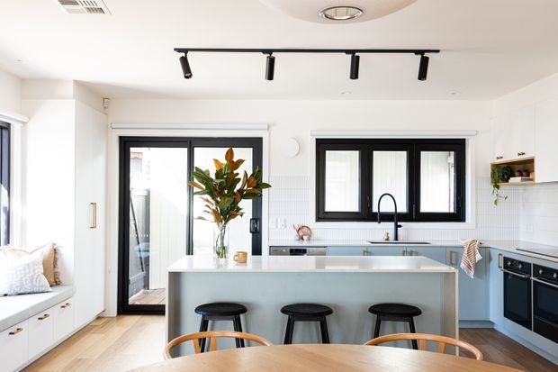 Bright contemporary kitchen with a large pale island, three black stools, light blue cabinetry, white tiled splashback, black-framed windows and doors, integrated appliances, timber flooring, and a round timber dining table in the foreground. A built-in window seat and open shelving add functionality and warmth.