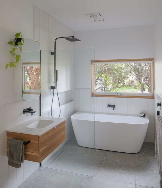 Bright contemporary bathroom featuring a freestanding white bathtub beneath a timber-framed window, a frameless glass shower enclosure with matte black fixtures, and a floating timber vanity with a white integrated basin. Large light-grey floor tiles and white vertically tiled walls create a clean, modern scheme. A wall mirror and hanging towel are visible, with greenery seen through the window.