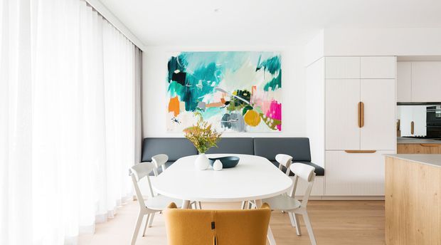 Bright contemporary dining area with a large white oval dining table, mixed white and mustard upholstered chairs, and a dark built-in bench seat beneath a colourful abstract artwork. Full-height sheer curtains line the left side, while white integrated cabinetry and a timber kitchen island are visible to the right. The space has light timber flooring and a clean, airy open-plan feel.