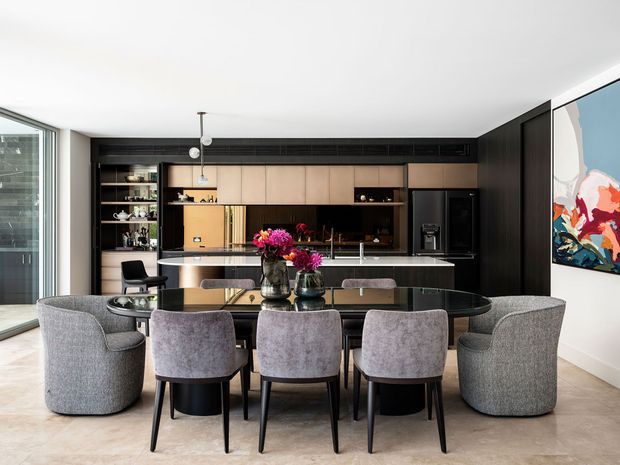 A sophisticated open-plan kitchen and dining area with a long dark dining table surrounded by upholstered grey dining chairs and armchairs. The kitchen features dark timber cabinetry, warm wood-toned upper cabinets, open shelving, a central island with a pale stone benchtop, integrated appliances, and a dramatic dark splashback. Large glazing provides natural light, while a colourful abstract artwork adds an accent to the space.