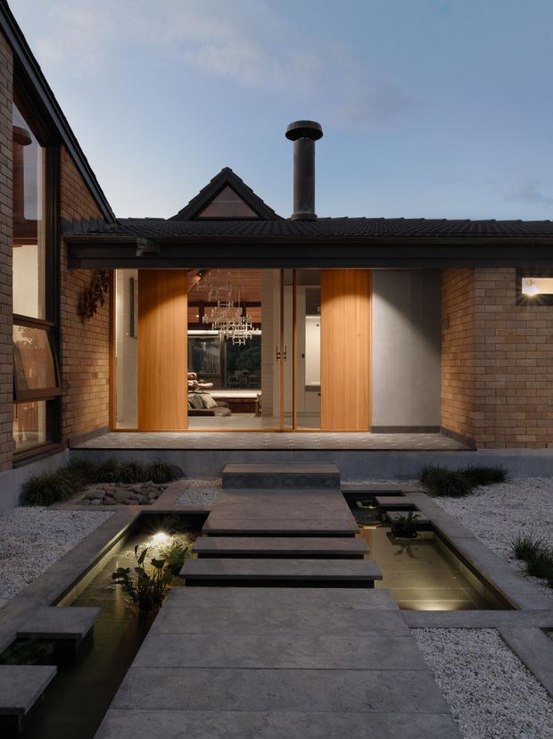 A dusk view into a carefully landscaped courtyard leading to a brick-clad residential entry. A broad concrete walkway crosses shallow rectangular reflecting pools via dark stepping slabs, with white gravel and low ornamental planting on either side. The house has dark tiled low-pitched roofs, exposed brick walls, timber-framed glazing, and large warm-toned timber entry doors. Through the open entry, a furnished interior and sculptural pendant light are visible. A prominent cylindrical metal chimney rises above the roof.
