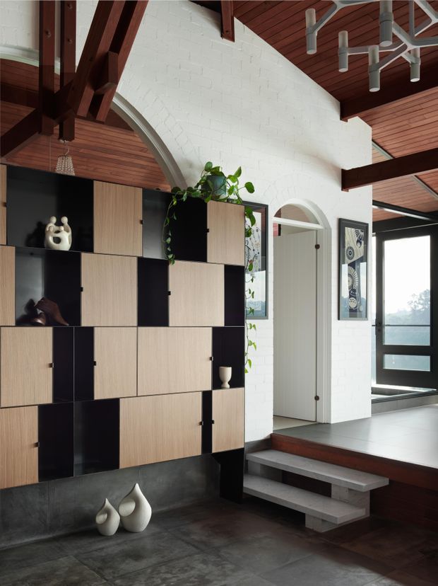 Interior view of a dramatic split-level entry and living area featuring a full-height white painted brick wall with arched openings, exposed dark timber beams, warm timber-lined ceilings, and a geometric built-in storage wall in alternating black and light timber panels. Concrete steps lead to a raised landing, with large dark-format floor tiles, framed artwork, sculptural decor, indoor plants, and expansive glazing at the entrance.