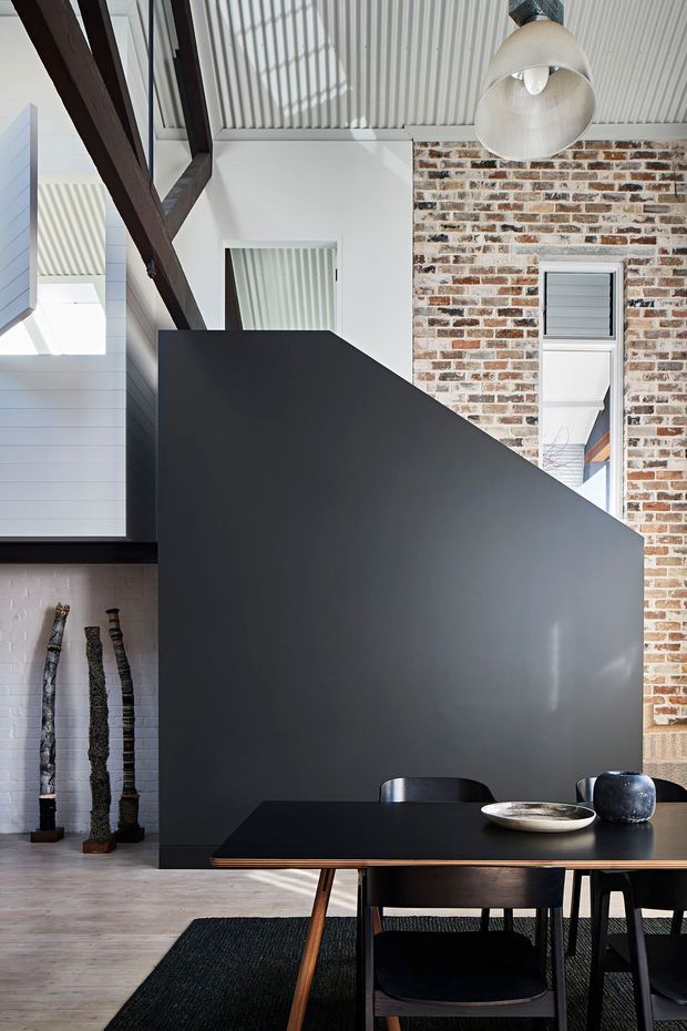 A dramatic contemporary dining area with a black dining table and chairs set against a tall matte charcoal partition wall. The space combines exposed aged brickwork, white painted wall cladding, dark timber structural beams, a ribbed white ceiling, and a large industrial pendant light. Decorative sculptural objects and a dark rug complete the refined monochromatic interior.