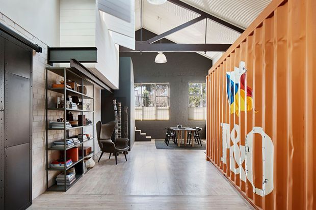 A bright industrial-style open-plan interior with a high pitched ceiling, exposed dark structural beams, white-painted masonry walls, and pale timber flooring. Open shelving and lounge chairs line the left side, while a vivid orange corrugated partition with colourful graphic artwork separates the dining area at the rear.