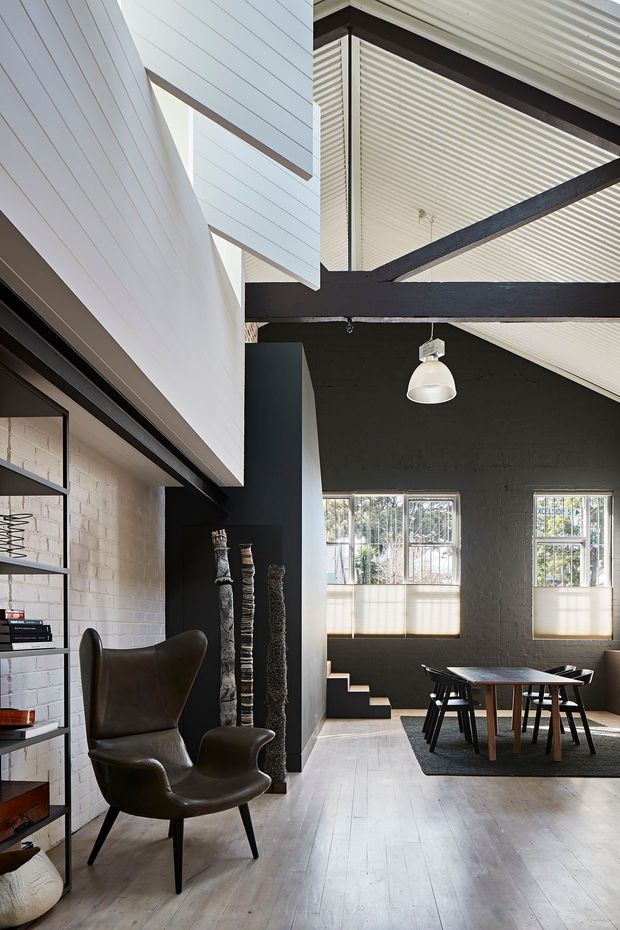 A tall, light-filled industrial-style interior combining a dining area and lounge. The space features exposed dark structural beams, a high pitched ceiling lined with white boards, black-painted brick walls, pale timber flooring, large multi-pane windows, open shelving, and a sculptural leather wingback chair. A dining table and chairs sit on a dark rug beneath a simple pendant light, with stairs leading to a raised area.