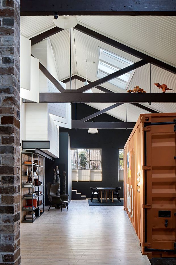 A dramatic double-height interior with exposed dark timber trusses, a white pitched ceiling with skylights, and dark painted walls. The space combines an open living and dining area, exposed brickwork, timber flooring, industrial shelving, and a striking orange shipping-container-style built-in structure. A stair leads down toward the dining area.
