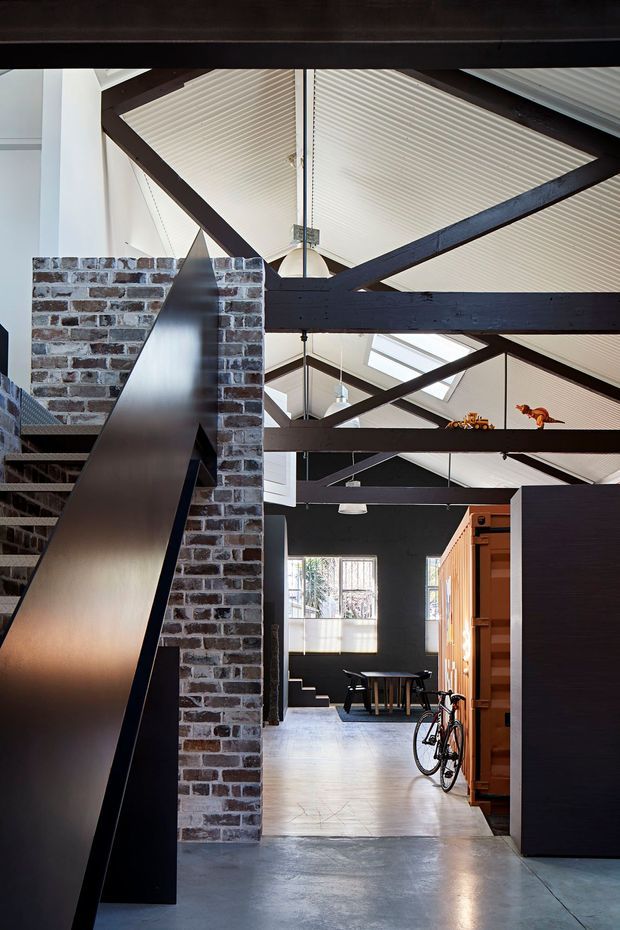 A dramatic industrial-style interior with exposed dark timber beams and trusses beneath a high pitched, ribbed ceiling with skylights. A raw brick masonry wall divides the space beside a sculptural dark metal stair or balustrade, while a dining area and timber joinery are visible deeper in the room. Polished concrete flooring, black walls, bicycles, and strong natural light reinforce the warehouse-conversion character.