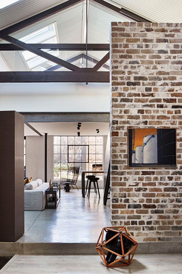 A spacious industrial-style loft interior with exposed dark timber or steel beams, a high glazed roof with skylights, polished concrete flooring, and a prominent exposed brick feature wall. The open-plan living and dining area includes a pale sofa, dark dining furniture, track lighting, and contemporary geometric decor.