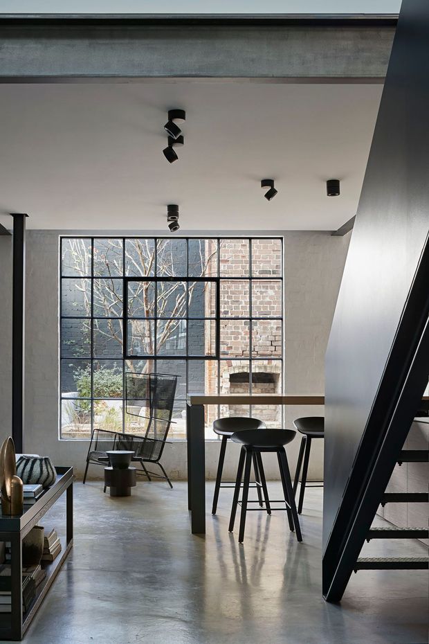A refined industrial-style open-plan interior with polished concrete flooring, a high white ceiling, exposed structural beams, and a large black steel-framed window overlooking a brick courtyard. A slim kitchen or bar counter with black stools occupies the centre, while a black staircase rises along the right side. A lounge chair, shelving, and small tables add restrained furnishings. The palette is predominantly grey, black, white, and brick brown, with strong natural light and clear architectural detailing.