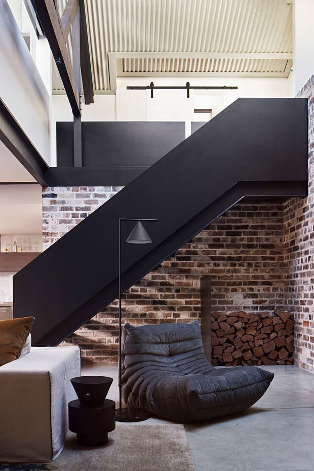 A dramatic contemporary living area beneath a black sculptural staircase. Exposed red and white brick walls, dark steel structural elements, a ribbed light ceiling, concrete flooring, and a built-in firewood niche create an industrial character. The room includes a sculptural lounge chair, sofa, and minimalist floor lamp.