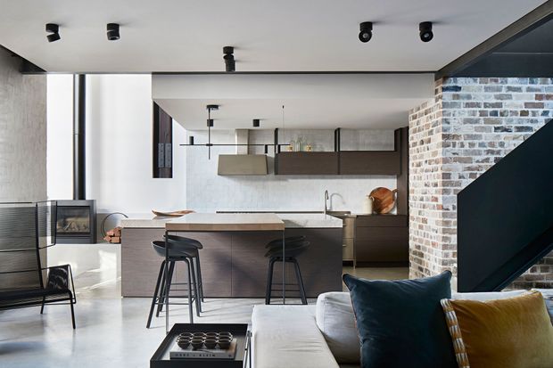Open-plan contemporary interior combining a kitchen, dining and living area. A large dark timber kitchen island with bar stools faces a pale upholstered sofa, while black-framed cabinetry, pendant lights, track lighting, exposed brickwork and a dark stair structure create a refined industrial character. A fireplace and floor-to-ceiling glazing are visible at the left.