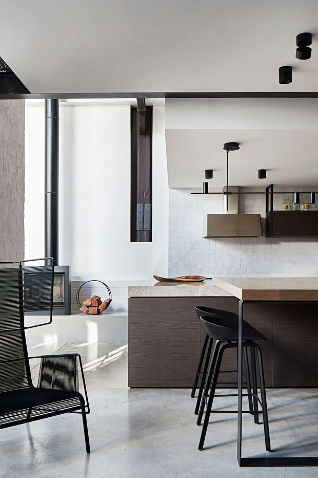 A refined contemporary kitchen in an open-plan interior, featuring a dark timber island with a pale stone benchtop, matching dark cabinetry, a tiled splashback, integrated rangehood, black bar stools, and exposed dark structural elements. Polished concrete flooring, black linear furniture, and minimalist black ceiling lighting create a high-contrast industrial-modern atmosphere.