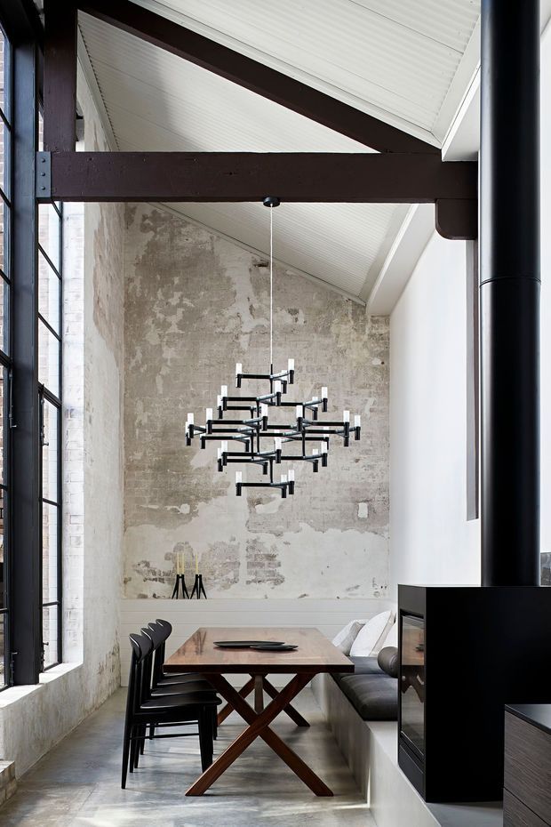 A dramatic dining area in a converted industrial-style interior, featuring a long timber trestle dining table, black dining chairs, built-in banquette seating, a sculptural black chandelier, exposed dark timber beams, tall black-framed glazing, a white vaulted paneled ceiling, and a distressed raw masonry wall. A black fireplace or joinery volume occupies the right side.