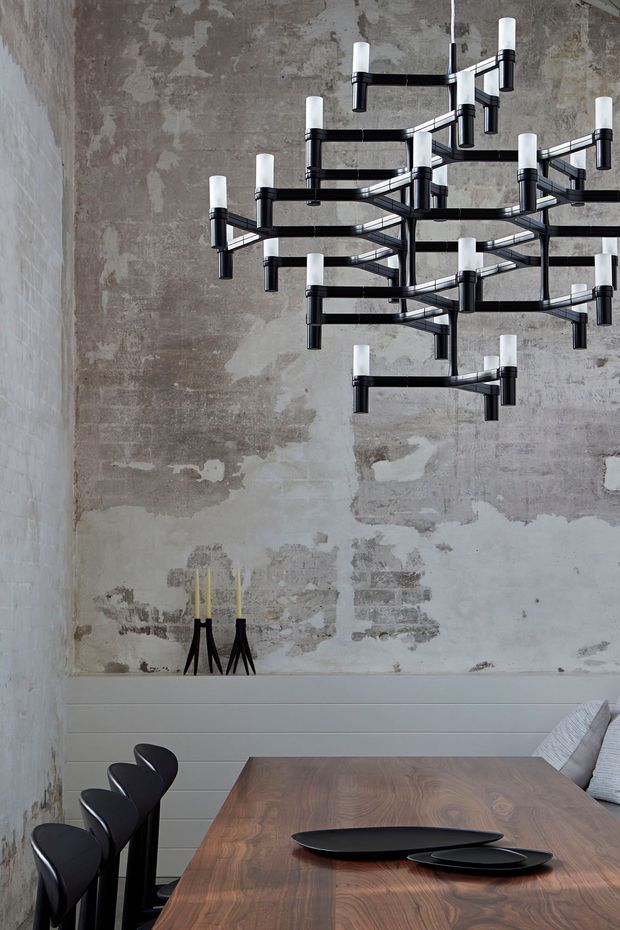 A refined dining room featuring a long solid timber dining table with black sculptural chairs, set against a textured raw plaster and exposed masonry wall. A large black geometric chandelier with white cylindrical bulbs dominates the upper portion of the room, while simple black-and-yellow decorative objects sit on a wall ledge. The space combines industrial architectural finishes with contemporary furnishings.