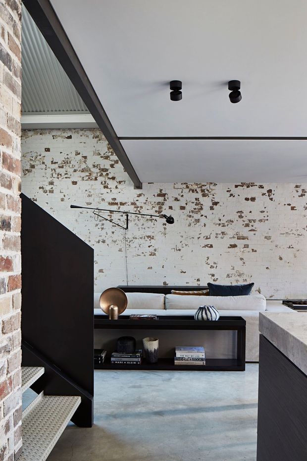 Interior view of an industrial-style open-plan living area adjoining a kitchen, featuring exposed white-painted brick walls with distressed surfaces, a polished concrete floor, dark built-in cabinetry and shelving, a light sofa, black track-style wall lighting, and a partially visible stair with textured metal treads. The ceiling is smooth and white with dark structural trim.