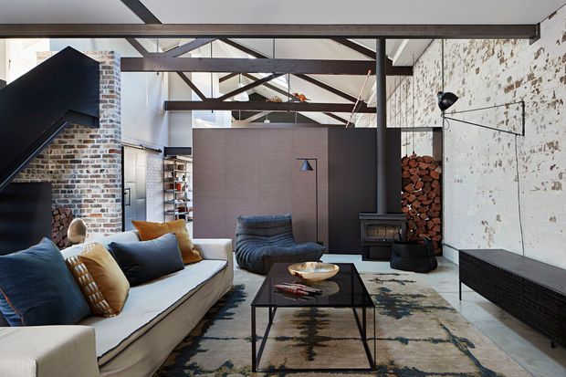 Industrial-style living room in a converted brick-and-concrete space, featuring exposed dark steel beams, a rough painted masonry wall, exposed brickwork, a black steel staircase, low-profile sectional seating, a sculptural lounge chair, a central coffee table, a freestanding wood-burning stove, and stacked firewood.