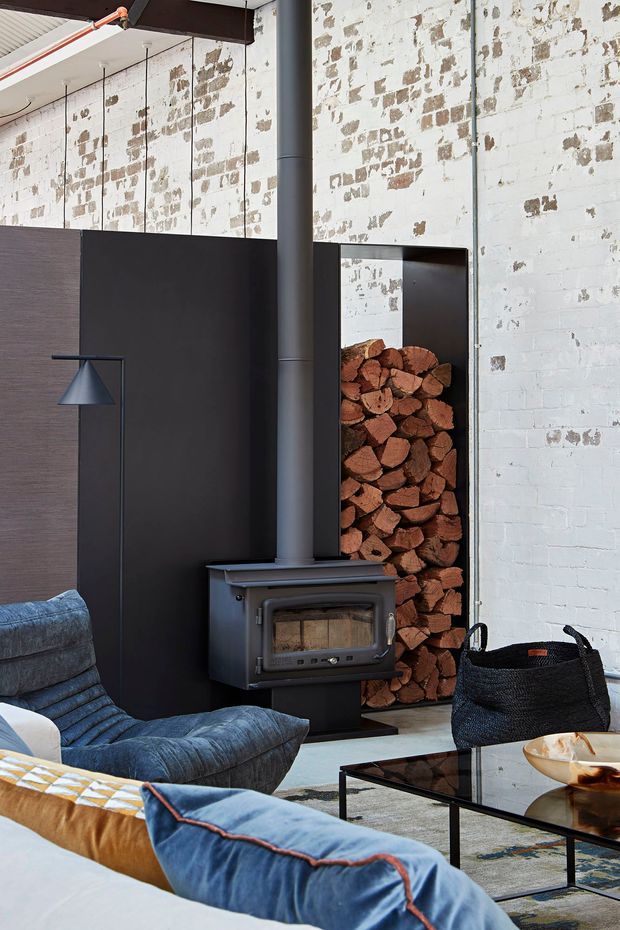 A stylish industrial living room featuring a freestanding black wood-burning stove with a tall flue, set against a distressed white-painted brick wall. A recessed niche stores split firewood beside dark wall panelling, while blue upholstered lounge seating and a black glass coffee table complete the room. Exposed structural beams and suspended lighting reinforce the warehouse-inspired character.